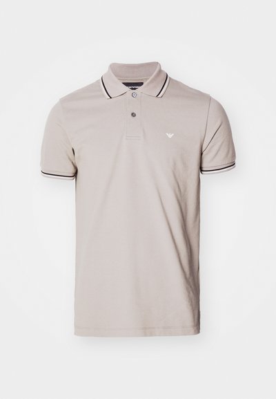 Polo bege claro feito de algodão, apresentando uma pala com três botões, gola e punhos de manga com riscas pretas e brancas, e um pequeno logótipo.