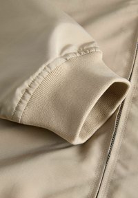 Μανίκι μπεζ ζακέτας με ριμπ λεπτομέρεια μανσετών που βρίσκεται δίπλα σε κλειστό μεταλλικό φερμουάρ πάνω σε λείο ύφασμα.