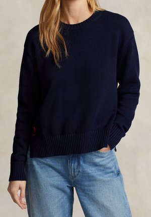Kobieta ubrana w granatowy dziergany sweter z prążkowanymi mankietami i dołem, w połączeniu z jasnoniebieskimi jeansami, jedną ręką w kieszeni.