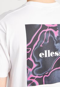Hvid bomulds-t-shirt med et stort abstrakt grafisk design i sort og pink på ryggen, med brandnavnet "ellesse" tydeligt vist.