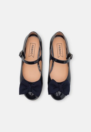 Une paire de chaussures Mary Jane en vernis noir avec des nœuds en tissu bleu marine et des brides de cheville, des semelles en beige et une étiquette de marque à l'intérieur.