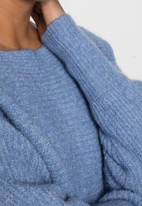 Pull en tricot bleu clair avec une finition texturée, doté de poignets côtelés et d'une coupe décontractée. Le tissu semble doux et chaud.