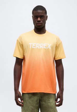 Mężczyzna ubrany w żółto-pomarańczową koszulkę TERREX i zielone kamuflażowe spodenki, stojący na tle jednolitego jasnego tła.