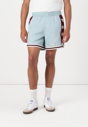 Mężczyzna w jasnoniebieskich i bordowych sportowych szortach z napisem „Hoop York City”, białe skarpetki sportowe i białe adidasy Adidas.