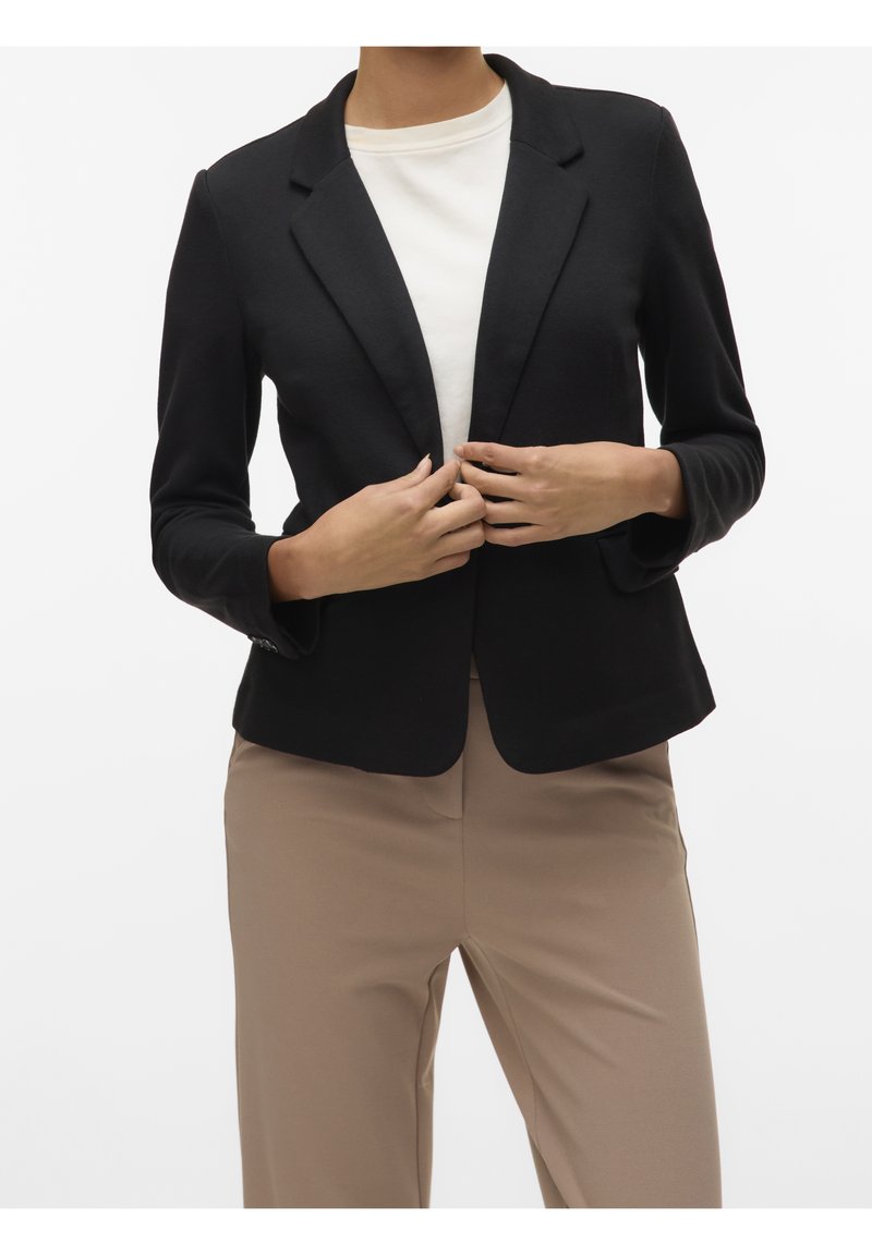 Zwarte blazer met een getailleerde pasvorm, scherpe revers en lange mouwen. Draagt over een lichtgekleurde top, gecombineerd met beige broek. Glad stoftextuur.