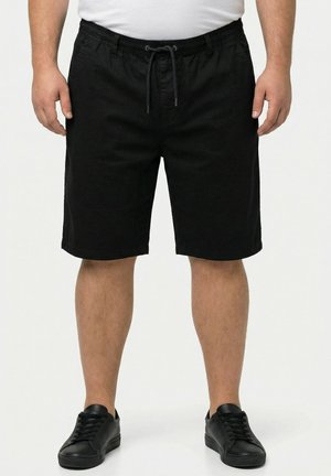 Mann trägt schwarze Shorts mit Kordelzug, schwarze Sneakers und ein weißes Hemd und steht mit den Händen entspannt seitlich am Körper.