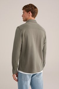 Hombre con cabello corto vestido con una camisa de manga larga color verde oliva y jeans claros, de espaldas contra un fondo beige liso.