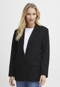 Črni blazer z enim gumbom, zarezane reverje in dvema sprednjima žepoma. Tkanina izgleda gladka, v kombinaciji z belo srajco pod njim.