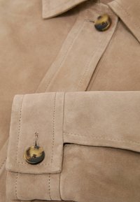Manche et poignet d'une veste en daim beige avec fermeture à bouton cousu et col assorti visibles en gros plan.
