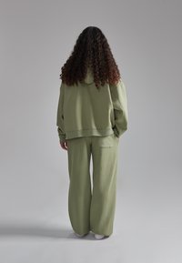 L'abbigliamento da casa verde oliva include una felpa oversize con cappuccio e pantaloni a gamba larga, realizzati in un morbido tessuto testurizzato con dettaglio della tasca posteriore.