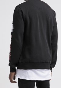 Sweatshirt nero con scollo rotondo, polsini e orlo a costine; presenta un motivo a zigzag rosa sulla manica sinistra. Indossato sopra una camicia bianca.