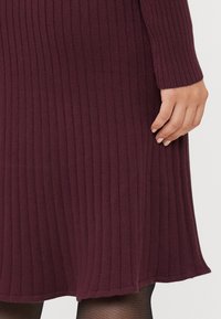 Burgundy ribbed knit dress with long sleeves, featuring a soft texture and a flared hem. Close-up view shows hand near the fabric.