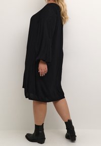 Robe noire knelt avec un tissu texturé, coupe ample et manches longues et bouffantes. Portée avec des bottines noires, montrant une silhouette lisse.