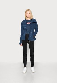 Navy zip-up jacket with unique hood design, fleece material, and side pockets, paired with dark jeans and white sneakers.