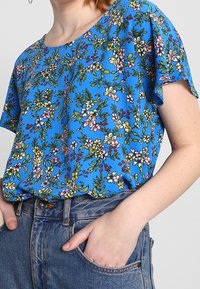 Modrý krátký rukáv top s květinovým vzorem v růžové, žluté a zelené barvě. Vyroben z měkké tkaniny, s uvolněným střihem a kulatým výstřihem.