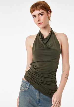 Jeune femme mince aux cheveux courts rouges portant un haut dos nu vert et un jean bleu, posant devant un fond blanc avec des tatouages visibles sur les bras.