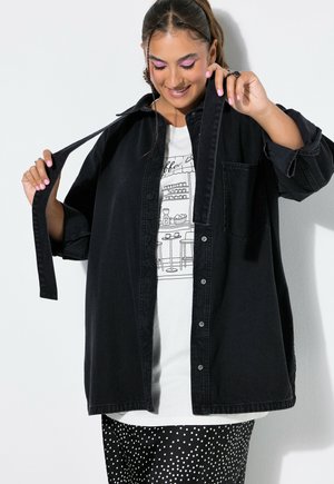 Vrouw die een losse zwarte blouse met knopen over een wit grafisch T-shirt en een zwarte polkadot rok aanpast, staand tegen een witte muur.