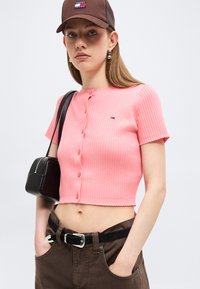 Νεαρή γυναίκα που φοράει καφέ καπέλο, ροζ κοντομάνικο ριμπ πουκάμισο με κουμπιά, καφέ παντελόνι με μαύρη ζώνη, κρατώντας μαύρη τσάντα ώμου.