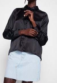 Blusa di raso nera con colletto, chiusura frontale a bottoni e maniche lunghe. Indossata con una gonna in denim chiaro con orlo tagliato.