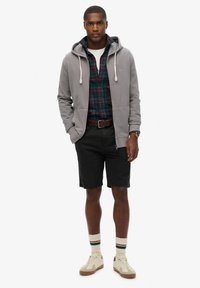 Gray hooded sweatshirt over a green and red plaid shirt, black shorts, cream sneakers, and striped socks with green, red, and cream accents.
