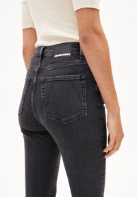 Jeans en denim noir avec un design ajusté, comportant deux poches arrière et une étiquette sur la taille. Tissu texturé avec de légers détails de décoloration.