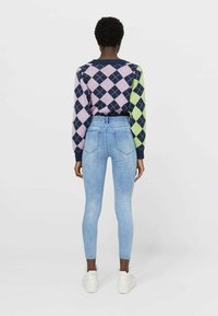 Suéter de punto colorido con patrón de rombos y mangas largas, combinado con vaqueros ajustados de talle alto en azul claro y zapatillas blancas.