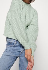 Jasnozielony, teksturowany sweter dziany z okrągłym dekoltem i dopasowanymi ściągaczami, zestawiony z niebieskimi dżinsami. Tkanina wydaje się miękka i przytulna.