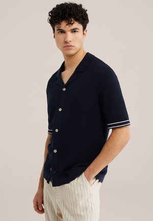 Fiatal férfi göndör hajjal, tengerészkék rövid ujjú kötött ingben fehér szegéllyel és világos színű, csíkos nadrágban, egyik kezét zsebében tartva áll.