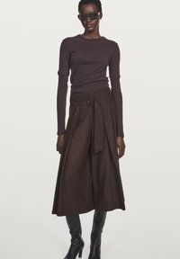 Donkerbruine top met lange mouwen gecombineerd met een broek met hoge taille en wijde pijpen. De broek heeft een ceintuurdetaillering. Model draagt zwarte zonnebril en knielange laarzen.