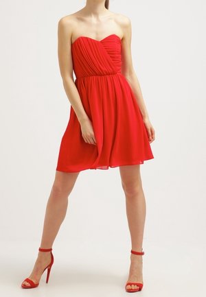 Femme portant une robe rouge sans bretelles jusqu'aux genoux avec un corsage plissé et des sandales rouges à talons hauts et brides à la cheville, debout, jambes écartées.