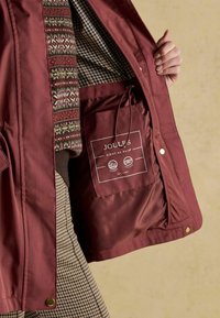 Burgundowa wodoodporna kurtka z wzorzystą podszewką i widoczną kieszenią na metkę. Posiada napy oraz gładką teksturę.