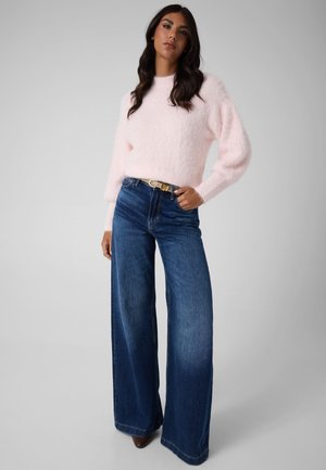 Femme portant un pull doux rose clair rentré dans un jean large taille haute bleu avec une ceinture noire et des chaussures marron, debout avec une main dans la poche.