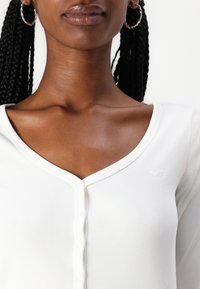 Haut long à manches blanches côtelé avec un décolleté en V et une fermeture à boutons. Présente un détail de logo subtil et une texture lisse.