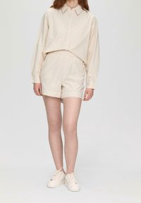 Camisa color crema con botones en la parte delantera y mangas largas, combinada con pantalones cortos a juego que tienen una cintura elástica. Lleva puestos unos zapatos deportivos claros.