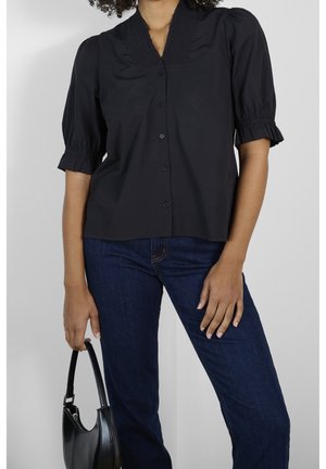 Femme portant un chemisier noir boutonné avec des manches bouffantes et un jean bleu foncé, tenant un petit sac à main noir à côté d'elle.