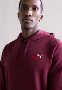 Homme portant un sweat à capuche maroon de Puma avec des cordons de serrage, regardant droit devant lui, sur un fond clair uni.