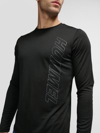 Homme portant un t-shirt noir à manches longues avec col rond, avec « HUMMEL » imprimé verticalement sur le devant, sur un fond uni.