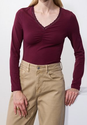 Femme portant un haut bordeaux ajusté à manches longues avec un décolleté en V en dentelle et un pantalon beige taille haute, debout les mains reposant près des hanches.
