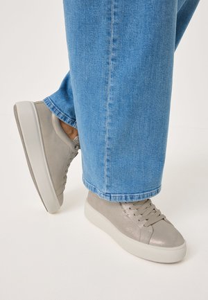 Sneaker in suede grigio con una finitura testurizzata e una suola in gomma bianca, abbinate a jeans a gambale largo blu. Le sneaker presentano lacci piatti e dettagli minimalisti.