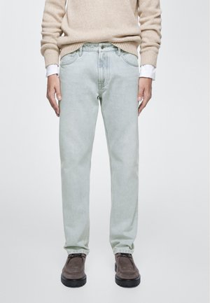 Uomo che indossa jeans azzurri, maglione beige, polsini di camicia bianca visibili e scarpe in camoscio marrone scuro, in piedi davanti a uno sfondo chiaro e uniforme.