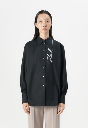 Camicia nera con bottoni, colletto, maniche lunghe e grafica bianca con firma sulla parte anteriore. Tessuto morbido, vestibilità rilassata.