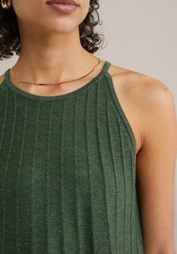 Zaļa ribota tank top blūze ar plānām siksniņām, izgatavota no teksturēta materiāla. Iezīmējas ar niansētu spīdumu un cieši pieguļošu dizainu. Redzama zelta kaklarota.