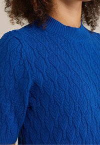 Blå kortärmad stickad tröja med ribbad hals och texturerat chevronmönster, bärs av en person med lockigt hår.