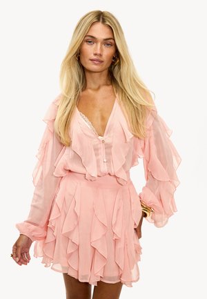 Robe en chiffon rose avec des couches froncées, un décolleté en V profond, des manches longues et une jupe évasée. Des accents en dentelle blanche embellissent le col.