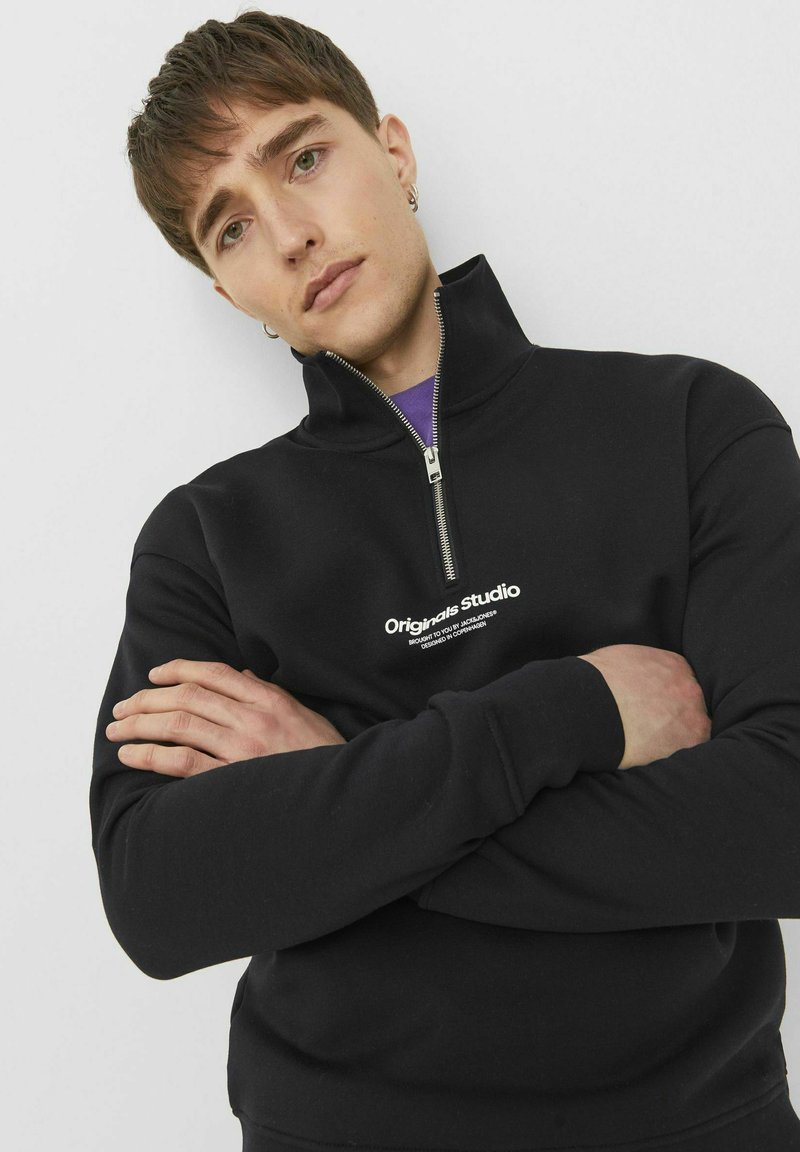 Zwarte rits-hoodie met hoge kraag, met een witte logo gecentreerd op de borst. Het materiaal lijkt zacht en gestructureerd.