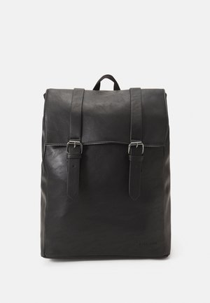 Sac à dos en cuir noir avec fermeture à rabat, deux sangles à boucles et une poignée supérieure, présenté sur un fond clair uni.