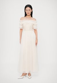 Biała suknia odkrywająca ramiona, z teksturowanym kwiatowym gorsetem i falbankowymi rękawami. Spływająca, plisowana spódnica sięga podłogi, zestawiona z białymi szpilkami.