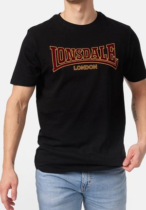 Mężczyzna w czarnej koszulce Lonsdale London i jasnoniebieskich dżinsach, trzymający lewy brzeg koszulki prawą ręką.