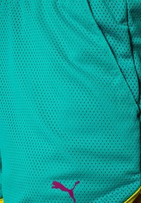 Short de sport bleu sarcelle avec de petites perforations, une poche latérale et un logo Puma violet près du bas avec une bordure jaune visible.