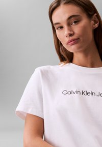 Fehér pamut póló kerek nyakkal és rövid ujjakkal, amelyen fekete betűkkel a "Calvin Klein Jeans" felirat olvasható a mellkason.